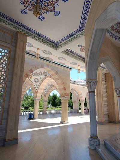 This media file captures an exterior view of a mosque, likely in Grozny, Russia, featuring its architectural details and surrounding environment. The scene is bathed in bright sunlight, suggesting it is daytime, and the weather appears to be clear with a bright blue sky visible through the arches.

The foreground is dominated by a marble-tiled floor and a series of arched colonnades. These arches are constructed with alternating light and reddish-brown stones, supported by large, ornate columns. The ceiling above the colonnades is a prominent feature, adorned with intricate geometric patterns and floral motifs in shades of blue, red, brown, and gold. Decorative lighting fixtures hang from the ceiling.

Beyond the colonnades, a well-maintained garden area is visible with lush green trees and a white balustrade. In the distance, the domes of the mosque's main structure are visible against the sky, with the characteristic crescent moon topping the central dome. A few people are seen in the middle ground, adding a sense of scale and human presence to the scene. One individual is dressed in a patterned long dress, while another person, partially obscured, is wearing dark clothing. A blue cleaning cart is also present on the tiled area.

The architecture showcases a blend of traditional Islamic design with possibly some modern influences. The rich ornamentation on the ceiling and the use of marble and stone suggest a grand and possibly recently constructed or well-maintained place of worship. The overall impression is one of serenity and grandeur, typical of religious sites. No visible text is present in the image.