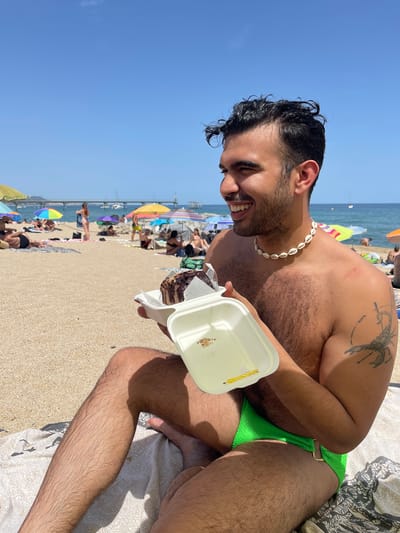 A man is sitting on a beach in Badalona, Spain, holding a container of cake. He is wearing green swim trunks and a shell necklace. The man has a tattoo of a scorpion on his right arm. In the background, there are beach umbrellas, people sunbathing, and the sea. A pier extends into the water in the distance. The sky is clear and blue.