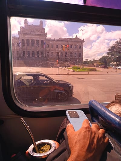 The image shows a person holding a light blue smartphone in their right hand, next to a blue bus seat bar. A mate gourd with a metal bombilla is situated to the left. Through a bus window, the Palacio Legislativo building in Montevideo, Uruguay, is visible, along with a street scene including a dark-colored car and traffic signals. The weather is sunny with clouds.