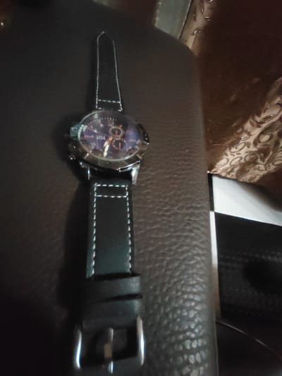 An overhead shot shows a black leather watch with white stitching on a dark brown, textured surface. The watch face is round with a dark blue dial and silver hour markers. Three sub-dials are visible on the face, and the brand name "RIBA" is printed on the dial. The watch band is made of black leather with white stitching and a silver buckle. The background is a mix of dark brown textured material and a patterned fabric. The scene does not provide any information about a specific location in Dorkovo, Bulgaria.