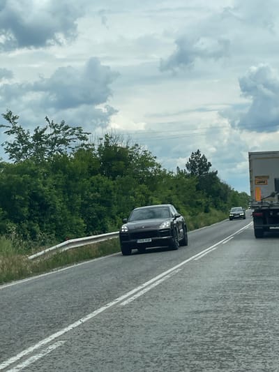 The image shows a road in Osenets, Bulgaria, with a black SUV car displaying the license plate "TR05 RGP". The car is driving on the right side of the road. In the distance, another car and a truck are visible. The background consists of trees and a cloudy sky.