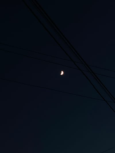 A crescent moon is visible in the dark night sky, partially obscured by several power lines. The lines run diagonally across the frame, creating a geometric pattern against the deep blue backdrop. The moon is small and emits a faint, warm glow. The scene appears to be an outdoor environment, likely in a city or town given the presence of power lines. The overall impression is one of quiet solitude and the vastness of the night sky.