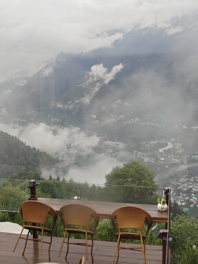 The image shows a view of a mountain valley with a town nestled within it, likely Prini, India, under a cloudy sky. In the foreground, there are three woven chairs and a wooden table on a wet wooden deck. The deck has a glass railing, and a small white planter with green plants sits on the table. Beyond the deck, lush green trees and dense vegetation cover the slopes of the mountains. The valley below is partially obscured by mist and clouds, with buildings of the town visible through the atmospheric haze. A river or road snakes through the valley. The overall scene suggests a scenic overlook, possibly at a hotel or restaurant, offering a panoramic view of the mountainous landscape.