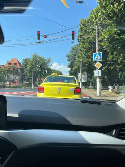 A yellow taxi cab is stopped at a traffic light in the city of Roman, Romania. The taxi is a Dacia Logan and has a "TAXI" sign on its roof. The traffic light is red, and there is a pedestrian crossing in front of the taxi. The image is taken from inside another vehicle. The weather is sunny.