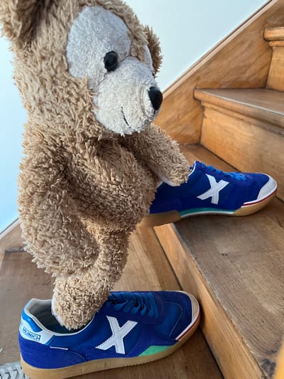 An indoor scene in Foetz, Luxembourg, depicts a light brown, shaggy-textured plush toy, resembling a bear, positioned as if ascending a wooden staircase. The plush toy, featuring a lighter muzzle and black button eyes, has its lower appendages inserted into two blue Munich brand athletic shoes. The shoes are characterized by blue suede-like uppers, blue laces, white toecaps, a prominent white 'X' logo on the side, and a multi-banded sole detailing in red, white, and green above a light brown gum sole. One shoe rests on a lower wooden stair tread, while the other is placed on the tread immediately above it. The staircase exhibits natural wood grain and signs of use. A light-toned wall or open space forms the background above the stairs.