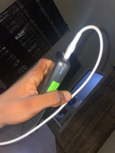 A close-up shot shows a person's hand holding a black portable charger. The charger is connected to a white cable, which is bent into a U-shape. There are four blue indicator lights on the charger, suggesting it is charging or has a certain battery level. A bright green sticker is visible on the side of the charger. The background is dark, with what appears to be a patterned curtain or blind on the left and a window with a blueish light on the right. The floor in the foreground is tiled. The scene suggests a domestic indoor setting, likely a bedroom or living area, in Malete, Nigeria.