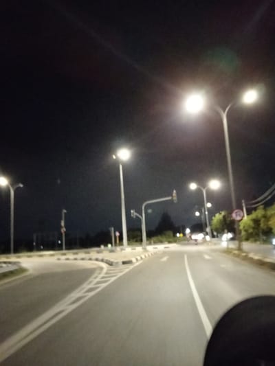 A nighttime view of a paved road, illuminated by streetlights. The road curves ahead, marked with white lines. A roundabout with black and white curbing is visible on the left. Traffic signals are present further down the road. The sky is dark. A portion of a vehicle or object is visible in the bottom right corner of the frame.