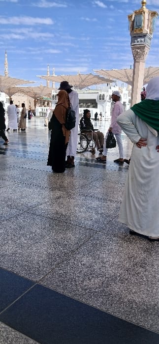 This image captures an outdoor scene at the Prophet's Mosque (Al-Masjid an-Nabawi) in Medina, Saudi Arabia, under a bright blue sky with scattered white clouds. The foreground is composed of a polished grey speckled tile floor with a darker blue rectangular border. In the mid-ground, a group of individuals stands on the paved area. On the far right, a person wearing a light-colored abaya and a green hijab stands with hands placed on their hips, facing away from the camera. To their left, a man in a pink thobe and a white skullcap holds a white plastic bag in his left hand, oriented towards a man seated in a manual wheelchair. The man in the wheelchair wears a dark jacket, dark pants, and a dark hat. To the immediate left of the wheelchair, a person in a black abaya and brown hijab wears a black backpack and stands beside a man in a white thobe and a dark hat. In the background, numerous individuals are dispersed across the expansive courtyard, some walking, others standing. Prominent architectural features include the white facade of the mosque buildings with arched windows, multiple slender minarets, and several large, light-colored retractable umbrellas. A decorative lamppost or pillar with golden accents is visible on the far right. The area is illuminated by daylight, casting shadows on the ground.