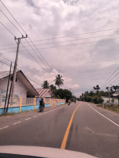 A wide-angle, eye-level shot captures a paved road stretching into the distance in a semi-rural setting in Ierhobglumpang, Indonesia, under a cloudy, overcast sky. The foreground reveals the top of a vehicle's dashboard, indicating the photo was taken from inside a moving car.
The asphalt road features a clear yellow solid line marking the center and white dashed lines on the shoulders. Three motorcycles are visible on the road. On the left shoulder, a rider in a black shirt and helmet is either stopped or moving slowly. Further ahead, two more motorcycles are seen, with one rider wearing a pink helmet.
On the left side of the road, a prominent concrete utility pole with multiple power lines stands against the sky. Behind it, a white house with a steep, light-colored tiled roof is partially obscured by a bright blue and yellow fence. Text visible on the fence includes "VISI" and "PENDING HARAPAN," along with part of a banner that reads "SMAN." Another house with a reddish-brown corrugated roof is nestled behind the first building, surrounded by lush tropical foliage.
Both sides of the road are lined with abundant greenery, dominated by numerous tall palm trees. Further down the road on the right, more residential structures and a white building with a satellite dish are visible. The time of day appears to be daytime, with diffused light due to the heavy cloud cover. The scene depicts typical daily activity along a local road in this Indonesian context.