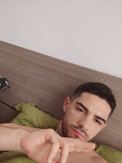 A medium shot depicts a man lying in bed. He is looking down at the camera with a slightly downward gaze. His hair is dark and neatly styled, and he has a trimmed beard and mustache. He is lying on a green pillow on top of a brown headboard. The upper portion of the frame is a white wall. The man's right arm and hand are visible in the foreground, angled towards the camera. The setting is likely an indoor bedroom. The location cannot be determined to be Fgura, Malta with the information given.