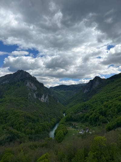 The image depicts the Tara River Canyon in Đurđevića Tara, Montenegro, with the river flowing through a deep gorge flanked by steep mountains covered in lush, green forest. On the left, a craggy, gray mountain rises, partially obscured by dramatic, billowing clouds. The sky is a mix of white clouds and patches of blue. There is a settlement with several small buildings and open areas near the river on the right side of the canyon.