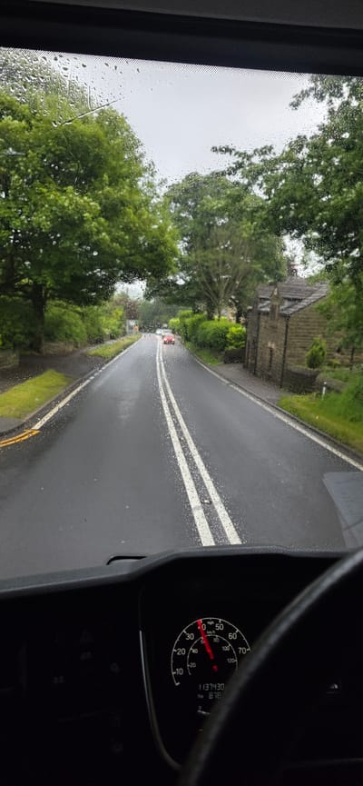 The image, taken from the driver's perspective inside a vehicle, shows a wet road with double white lines, indicating a two-lane road. Raindrops are visible on the windshield. Lush green trees line both sides of the road, and a stone building with a pitched roof is situated on the right. In the distance, two red vehicles are visible. The vehicle's dashboard is in the foreground, displaying a speedometer indicating approximately 40 mph. The odometer reads 1137430, and the trip meter reads 878. The image appears to have been taken in Barnsley, United Kingdom, given the context of the request.