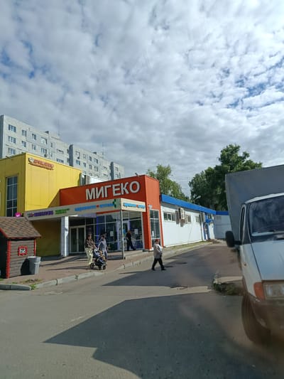 A street-level, medium shot captures a sunny day in what appears to be a commercial area in Orekhovo-Zuyevo, Russia. The sky is partly cloudy, with large white clouds dominating the upper portion of the frame. In the foreground, a grey asphalt road is visible, casting a long shadow to the right, suggesting the sun is high in the sky, likely mid-morning or early afternoon.
To the left of the road, a paved walkway leads to a row of storefronts. The most prominent building is a bright orange structure with a large, white "МИГЕКО" sign emblazoned across its upper facade, indicating a commercial establishment, possibly a supermarket or shopping center. To the left of the orange building, a bright yellow structure houses "ВЯЗАНКО", a store that appears to specialize in knitwear or similar textile products, based on its logo which depicts a steaming bowl. Adjacent to the yellow building, a small, whimsical wooden playhouse with red trim and a depiction of a teddy bear adds a touch of local character.
In front of the stores, several people are engaged in everyday activities. A woman is pushing a stroller with a child in it, walking along the sidewalk. Another woman is seen walking towards the right of the frame. A few individuals are visible inside the "МИГЕКО" store through its glass facade. A white delivery truck, partially visible on the far right, suggests ongoing commerce or deliveries.
The scene depicts a typical day in a Russian town, with a mix of residential buildings in the background, commercial establishments, and people going about their daily routines under a bright, cloudy sky. The visible text includes "МИГЕКО", "ВЯЗАНКО", and smaller signs that are difficult to read. The overall mood is one of casual urban life.