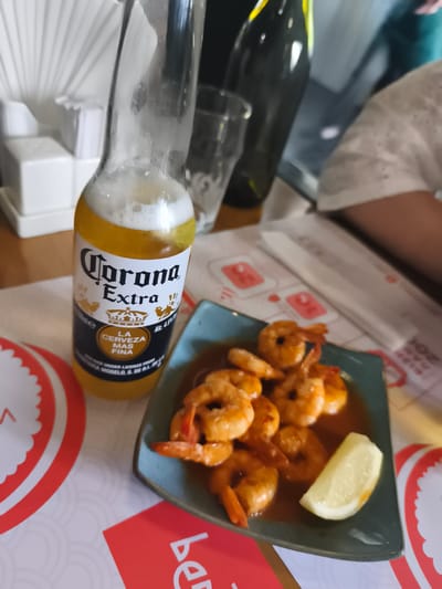 This media file depicts a close-up view of a table setting with food and drinks. The primary subjects are a bottle of Corona Extra beer and a plate of glistening shrimp. The beer bottle, partially filled and with a foamy head, is prominently positioned in the foreground, its label clearly visible with "Corona Extra" and the crown logo. To the right, a blue-green plate holds several cooked shrimp coated in a glossy sauce, accompanied by a wedge of lemon.

In the background, other objects suggest a dining environment. A dark bottle, possibly wine, stands behind the beer. A clear glass and a white napkin dispenser are also visible. The table appears to be covered with placemats that have red and white circular patterns and some pink graphic elements. The lighting suggests an indoor setting, possibly a restaurant or bar, during the daytime. The overall mood is relaxed and inviting, implying a meal or a casual gathering.

The visible text on the beer bottle includes "Corona Extra," "LA CERVEZA MAS FINA," and "Alc. 4.5%." There are also some dates and other smaller text elements on the bottle that are not entirely legible. The placemats also have some text, including what appears to be "be" in large white letters on a red background at the bottom right.

While the image provides no explicit information about the specific time of day or weather, the indoor setting and the presence of food and drink suggest a meal or social occasion. There are no visible people in the frame, but the context implies their presence at the table. The location context provided is Kryzhanivka, Ukraine.