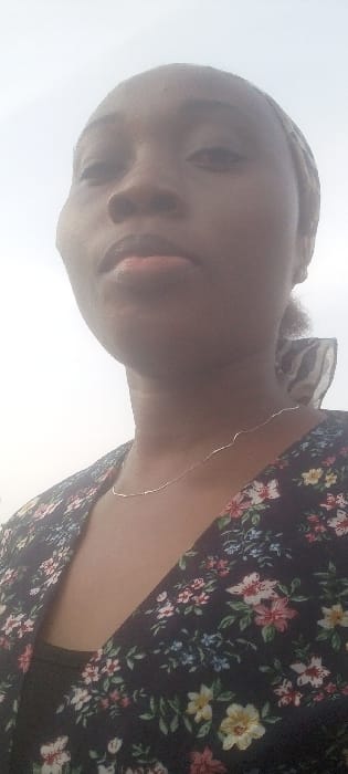 A woman is looking up at the camera from a low angle. She is wearing a black dress with a floral print. She has on a delicate silver necklace. The background is a bright sky. The woman's skin tone is brown, and she has full lips with red lipstick. Her eyes are partially closed. The image has a slightly blurry quality, possibly due to the lighting conditions or camera settings.