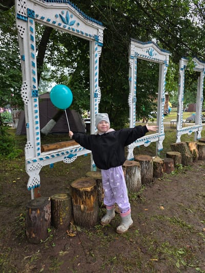 A young child stands outdoors, holding a teal balloon on a stick. The child is wearing a black jacket, light purple pants with white heart patterns, and grey boots. They are positioned in front of three ornate, white and blue decorative frames, which appear to be part of an outdoor installation. Behind the frames, a large, dark tent is visible, along with trees and foliage. The ground is muddy and covered with grass and fallen leaves, suggesting a natural or park-like setting. The overall scene appears to be in a rural location, possibly the village of Staroye Bygi, Russia, given the context.