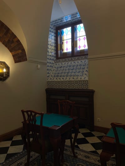 The image shows a room with antique furnishings, likely in a historic building. A dark wooden table with a teal surface, resembling a card or gaming table, is in the foreground, surrounded by wooden chairs. To the right, another table with a similar teal surface is partially visible. The floor is a checkerboard pattern of black and white tiles, with an oriental rug in front of the table.
The wall behind the table is adorned with blue and white decorative tiles, and above them, a stained-glass window with geometric patterns in shades of blue, yellow, and pink is visible. A wooden cabinet with intricate carvings is situated below the window. To the left of the frame, a brick archway is visible, with a sconce holding a faux candle mounted on the wall.
This scene is located within the Catherine Palace in Strelna, Russia, which is part of the Peterhof State Museum-Reserve. The palace is known for its opulent interiors and historical significance, serving as a summer residence for Russian royalty. The presence of antique furniture, decorative tiles, and stained glass suggests a room that retains its original historical character.