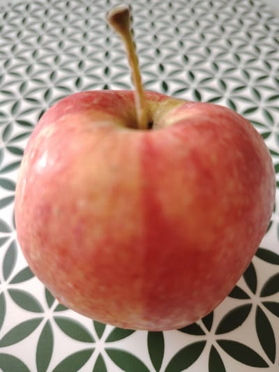 A close-up, eye-level, slightly angled shot captures a single, ripe red apple placed on a surface with a repeating pattern of green and white geometric shapes. The apple is positioned in the center of the frame and is slightly out of focus, with its stem visible extending upwards. The surface appears to be a tabletop or countertop. The image does not contain any information about the city of Кривец, Russia.