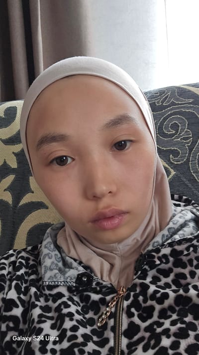 A close-up, selfie-style portrait shows a person wearing a beige hijab and a black and white leopard-print hooded jacket with a gold zipper. The person's face is centered in the frame, with brown eyes looking directly at the camera. Their lips are slightly parted, and their skin appears smooth. The background consists of a textured couch with a dark gray and white pattern. To the left, a portion of brown curtains is visible. The image is marked with "Galaxy S24 Ultra" in the lower-left corner. The environment suggests an indoor setting, possibly a home or a comfortable seating area. No specific location within Tilekmat, Kyrgyzstan, can be identified from the visual information provided.