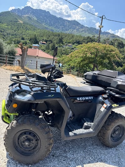 A black Segway ATV is parked on a gravel surface in front of a fence. The ATV has a cargo rack on the front and a storage box on the back. The handlebars are visible, along with mirrors and handguards. In the background, there is a building with a red-tiled roof, trees, and a mountainous landscape under a blue sky with scattered clouds. The location is identified as Rachoni, Greece.