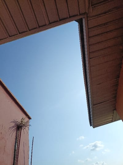 The image presents a striking view looking directly upwards towards a clear, bright blue sky, framed by various architectural elements. The perspective is from a low angle, suggesting the viewer is looking up from between buildings or structures.

The sky occupies the majority of the frame and is a vibrant, uniform shade of light blue, indicating a sunny day. In the bottom-left corner of the sky, a few wispy white cumulus clouds are visible, adding a touch of texture to the otherwise expansive blue.

Framing the sky are sections of what appear to be building roofs or walls. In the upper-left portion of the image, a flat, reddish-brown structure cuts diagonally across the corner. This surface appears somewhat worn or aged. From a crack or crevice near its edge, a small, hardy plant with spiky leaves is growing, casting a distinct shadow onto the reddish-brown surface, highlighting the strong sunlight. A thin, dark, linear object, possibly a wire or a small antenna, extends downwards from this structure into the sky.

On the right side of the image, and extending along the bottom-right and slightly into the bottom-center, is a different structure. This element is predominantly dark brown and features prominent horizontal corrugations or ribs, characteristic of corrugated metal roofing or siding. The lines of this material run horizontally, creating a textured pattern. The edge of this structure forms a sharp, dark line against the bright sky.

Another section of a similar dark brown, corrugated structure is visible in the bottom-middle and bottom-right, forming an angled line against the sky. Just below its uppermost edge, a dark, recessed channel runs horizontally, possibly indicating a gutter system or a trim detail.

The overall impression is one of geometric abstraction, with the architectural lines sharply contrasting with the soft expanse of the sky. The bright lighting suggests midday sun. There are no people or discernible activities taking place, and no text is visible in the image. The scene conveys a sense of quiet observation of the sky framed by man-made structures.