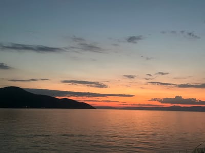 A wide shot captures a serene sunset over the Aegean Sea, likely in the vicinity of Thassos, Greece. The sky transitions from a pale blue at the top to a vibrant orange and yellow hues closer to the horizon, indicative of dusk. Several dark, silhouetted clouds are scattered across the sky, some tinged with the orange glow of the setting sun.
A large, dark mountain range dominates the left side of the frame, its peak casting a shadow over the water. The sea itself is calm, reflecting the warm colors of the sky in gentle ripples. In the distance, along the coastline, faint lights suggest a settlement. A small, dark boat is visible on the water to the right of the mountain. The foreground shows a sliver of a white railing, possibly from the viewpoint of a ferry or boat. No people are visible in the frame. The overall impression is one of tranquility and natural beauty at the end of the day.