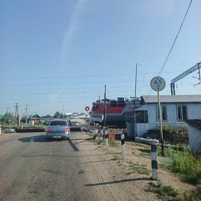 The image depicts a railway crossing in Kambarka, Russia, on a clear, sunny day. A red and grey electric locomotive is stopped at the crossing, with its red signal light illuminated. A blue sedan is stopped behind the locomotive on the road. A circular sign indicating a height limit of 4.5 meters is visible. The crossing is equipped with barriers and striped marker posts. The environment includes a small, light-colored building to the right of the tracks, with vegetation and debris piled nearby. The road surface appears to be asphalt. In the background, there are several houses with pitched roofs, power lines, and trees, suggesting a semi-urban or rural setting.