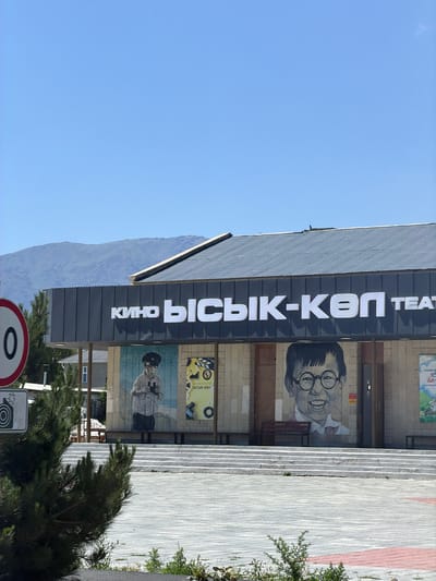 The media file shows the exterior of a cinema and theater building in Balykchy, Kyrgyzstan. The building's facade features a prominent sign with white lettering against a dark background, reading "Кино ЫСЫК-KON TEAT" in Cyrillic and Latin characters, suggesting the name "Ysyk-Kol Cinema-Theater".
Below the main sign, there are several murals. One depicts a black and white portrait of a person with glasses and a distinctive hairstyle. Another mural shows a person in a hat and uniform holding binoculars. A smaller poster above this mural has text that seems to be advertising.
The building is situated next to a paved walkway with a patterned surface. To the left of the building, a steep, blue-toned mountain range is visible under a clear blue sky, indicating a daytime scene. Green trees and shrubs are present in the foreground and to the right of the building, suggesting an outdoor setting in a populated area. In the lower part of the image, a traffic sign with a red circle and a number "0" is partially visible, along with another sign indicating a camera symbol. The overall impression is a functional building serving as a cultural hub within a town that borders natural landscapes.