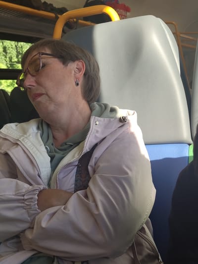 An older woman with short brown hair, glasses, and earrings is sleeping on a bus. She is wearing a light pink jacket over a green hooded sweatshirt. Her arms are crossed. The background shows a yellow handrail and a light blue and grey seat. The woman is in Zolotovo, Russia.