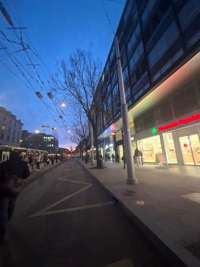 This image captures an urban street scene at dusk or early evening, rotated ninety degrees counter-clockwise. The sky on the left is a deep, dark blue, against which the bare branches of a tree are silhouetted. A large, modern building occupies the upper portion of the frame (left side when viewing upright), featuring an extensive glass and metal canopy that extends over a sidewalk. A tall, light-colored pole stands vertically, supporting this overhead structure.
On the building's facade, bright red lettering clearly reads "Pharmacie Populaire," accompanied by a prominent green cross symbol, identifying it as a pharmacy. The building's large windows display brightly lit interiors, suggesting a commercial establishment.
The right side of the image depicts a wide, dark paved area, possibly a street or a large pedestrian plaza, with a distinct yellow zigzag line painted on its surface. Numerous streetlights and building lights illuminate the background, creating a warm glow. Several people are visible in the distance, walking along what appears to be a busy street or public space. Closer to the foreground on the right, a person in a dark jacket is seen from behind. Overhead, multiple cables with decorative or functional lights are strung across the scene, contributing to the evening ambiance.
The visible French text "Pharmacie Populaire" strongly suggests a location in a French-speaking region, aligning with the provided context of Genève, Switzerland. The scene depicts a typical, calm evening in a European city.