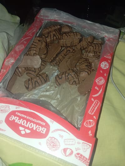 The image shows a box of chocolates from the "Belogorye Confectionery Factory". The chocolates are individually wrapped and appear to be a type of wafer cookie covered in chocolate with chocolate drizzle on top. Some of the chocolates are broken. The box is red and white and has various decorative drawings on it, including what appear to be baked goods and wheat stalks. The box is sitting on a yellow surface, possibly a bed or a couch, with a white cloth to the left. The lighting is dim, suggesting the photo was taken indoors at night or in a poorly lit room. There are no people visible in the image. This particular confectionery factory is located in the city of Rahachow, Belarus.