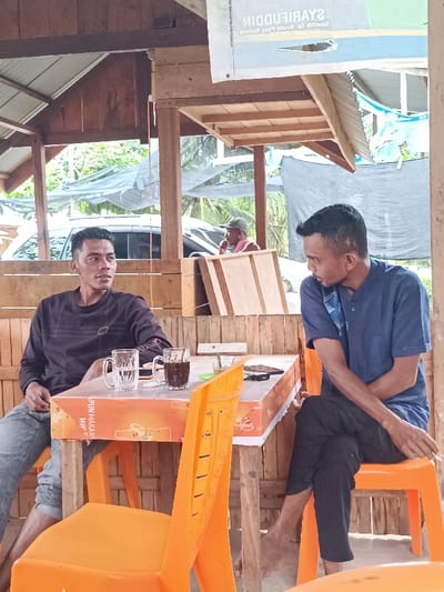 Two men sit at a table with drinks, engaged in conversation. The man on the left wears a dark long-sleeved shirt and jeans, while the man on the right dons a blue button-down shirt and dark pants. They are seated on orange plastic chairs at a table covered with an advertisement. A third, older man sits in the background, partially obscured by stacked wooden panels. The scene is outdoors under a simple shelter structure with a corrugated metal roof. A vehicle can be seen in the background.