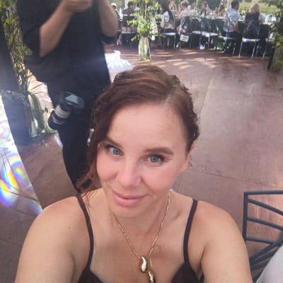 The image is a selfie taken by a woman with brown hair and blue eyes. She is wearing a dark-colored top and a gold necklace with two teardrop-shaped pendants. Her expression is a slight smile. In the background, there are people seated at tables, suggesting an outdoor event like a wedding or party. The setting appears to be a park or a garden with trees and a vase of greenery on a table. There is also a photographer in the background holding a camera. The ground is paved. The city name "Svetloe, Russia" is mentioned in the prompt, but there are no visual cues in the image to confirm this location.