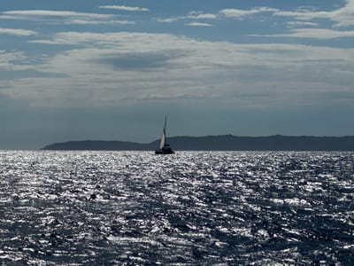 The image depicts a vast expanse of choppy, sun-dappled dark blue water under a cloudy sky. In the distance, a silhouetted catamaran with its sails raised is visible, navigating the waters. Behind the catamaran, a dark, hazy silhouette of a coastline or island forms the horizon. The scene is illuminated by sunlight reflecting off the water's surface, creating a glittering effect. The overall impression is one of a calm, open-sea environment. The location is identified as Bormes-les-Mimosas, France.
