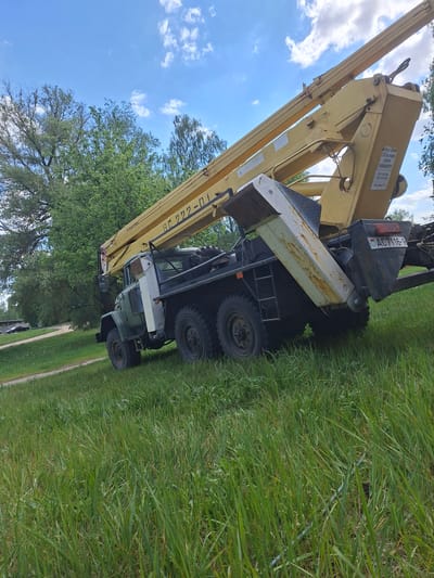 The image shows a yellow truck crane parked on grass, possibly in Zhabinka, Belarus, as indicated by the Belarusian license plate "AC7116-1". The boom is extended towards the sky, partially obscuring the view of trees and the partly cloudy sky. The truck has multiple wheels and appears to be heavy-duty. Text on the boom reads "БF 222-01" and provides safety instructions.