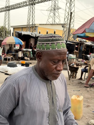 An adult Black male is depicted in a semi-close-up, looking downwards and to his right, in an outdoor market setting in Jakande, Nigeria. He wears a gray, vertically striped tunic with intricate gray embroidery around the placket and neckline, and a green and white embroidered kufi cap featuring leaf-like patterns. His short beard is dark with some gray.
Behind him, an active market scene unfolds under an overcast sky. To the left, a stall with a striped blue, white, and tan umbrella features white basins and stacked bags, with multiple individuals partially visible. To the right, another stall with a reddish tarp roof is present, with a person seated and a dark pot on a wooden stand. A yellow plastic jerrycan rests on the dusty, unpaved ground in the mid-right. In the far background, two large metal lattice electrical transmission towers rise above a yellow-walled building with a gray roof, indicating a proximity to power infrastructure.