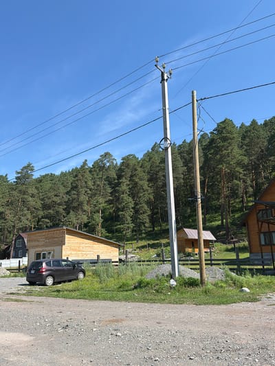 In Cheposh, Russia, a gravel driveway leads past a dark-colored hatchback car parked on grassy terrain. To the left, a modern wooden building with a beige base and a light brown wooden upper section stands under a clear blue sky. Behind the car and building, a dense forest of tall green pine trees covers a hill. Power lines stretch across the frame, supported by two utility poles: one concrete and one wooden. A small wooden structure with a metal roof is visible further up the hill, nestled among the trees, and part of a wooden fence can be seen.