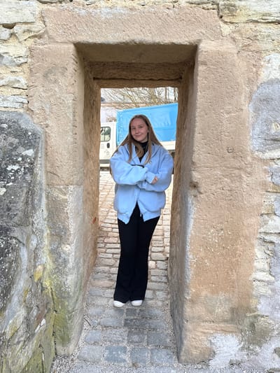 A young adult female stands centrally positioned within a narrow, weathered stone archway in Weißenburg in Bayern, Germany. The archway is constructed from large, light-colored, rough-hewn stone blocks, exhibiting visible signs of age, including a square indentation on the right jamb and moss/lichen growth on the left jamb. The subject possesses long, light brown hair and is dressed in a light blue, oversized, zip-up hoodie worn over a black turtleneck top, black trousers, and white athletic shoes. Her arms are crossed at the chest, and she looks directly at the camera with a slight smile. The ground beneath her within the passage is paved with irregularly shaped grey cobblestones. Through the archway, a white panel van or truck with a prominent bright blue tarp covering its cargo area is visible, parked on a paved surface. Further in the background, bare trees are visible against a light sky.