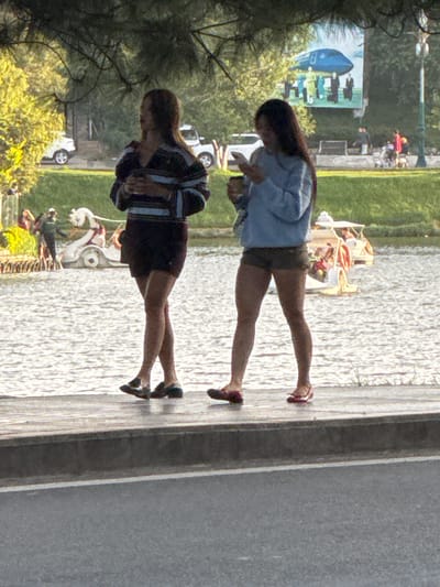 Two young women walk along a paved path next to Xuan Huong Lake in Da Lat, Vietnam. The woman on the left wears a striped long-sleeved shirt and dark shorts, holding a cup in her hand. The woman on the right is dressed in a light blue long-sleeved shirt and dark shorts, and is looking at her phone. The lake's surface is visible behind them, reflecting sunlight. In the background, there are trees, a white vehicle, and a billboard with an image of an airplane. Further in the distance, people and a paddle boat are visible. The scene appears to be during daylight.
