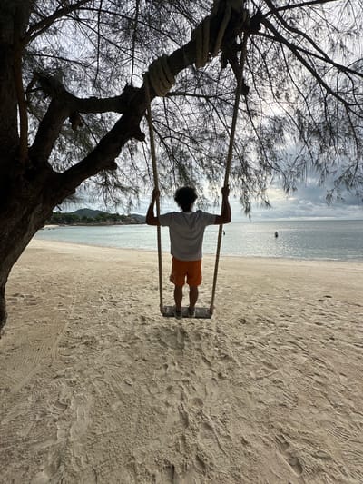A person is seen from behind, sitting on a swing suspended from a tree by ropes. The person is wearing an orange and white striped shirt and orange shorts. They are facing the ocean, with a sandy beach in the foreground and lush green hills in the distance. The sky is overcast with dark clouds, and the sea is calm. The scene appears to be a peaceful, tropical setting on Ko Pha-ngan District, Thailand. There are two other people in the water, further out in the ocean. The sand is covered with footprints and tire tracks.