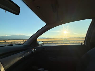 The image depicts the interior of a car, looking out the passenger side window towards a landscape bathed in the golden light of sunset. The scene captures a vast, golden field stretching towards a distant city skyline. The city, which is likely Kazichene, Bulgaria, is silhouetted against the warm sky, with numerous tall industrial structures, possibly factories or power plants, dotting the horizon. A highway runs through the foreground, with a visible railing.

Inside the car, the dashboard and door panel are visible. A small, decorative item featuring tiny purple flowers in a miniature vase is attached to the door panel. A rearview mirror is visible at the top of the windshield. The sun, low in the sky, casts a bright glare through the windshield. The overall impression is one of a serene, end-of-day journey, with the natural beauty of the Bulgarian countryside juxtaposed with the urban sprawl of the city.
