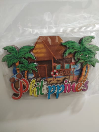 This is a close-up, eye-level, full shot of a souvenir magnet shaped like a tropical beach scene. The magnet depicts a thatched-roof hut on stilts, surrounded by palm trees. In the foreground, the word "Philippines" is written in colorful, bubbly letters. The magnet is enclosed in clear plastic packaging, which reflects some light. The background is a plain white surface. There are no people or animals visible in the image, and no specific location in Tervel, Bulgaria is identifiable from the scene.