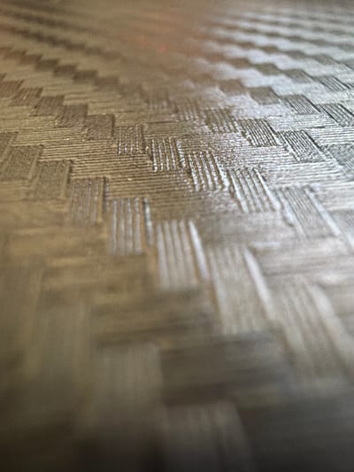 The image is a close-up, angled shot of a woven carbon fiber textured surface. The texture consists of alternating bands of parallel lines, creating a diagonal weave pattern. Some areas of the surface appear brighter, indicating light reflection. The overall color palette is muted browns and grays. There are no people, animals, or distinct objects visible in the image. The background is out of focus and appears to be a continuation of the same textured surface, fading into darkness at the bottom of the frame. The image does not contain any discernible features that would allow for location identification, including within Grapevine, United States of America.