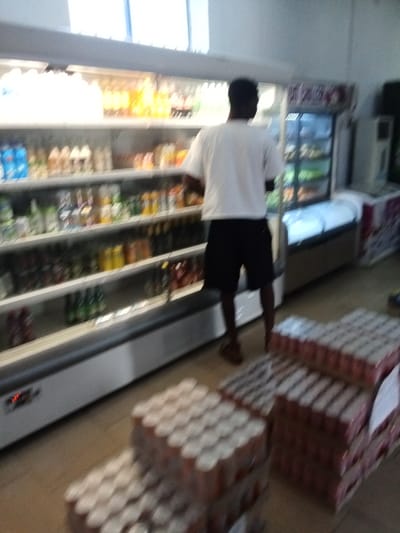 The image shows the interior of a shop located in Sapele, Nigeria. It appears to be a grocery store. There is one man, dressed in a white t-shirt and black shorts, in the photo. He is looking into the open door of a refrigerated display case filled with various beverages. There are stacks of boxes of canned goods in the foreground. The boxes are stacked in front of another refrigerated display case. The shop appears to be well-lit and relatively clean. The floor is tiled. The walls are painted white. There is a large window in the background, but its view is obscured. There are a couple of other display cases in the background. The image is out of focus.