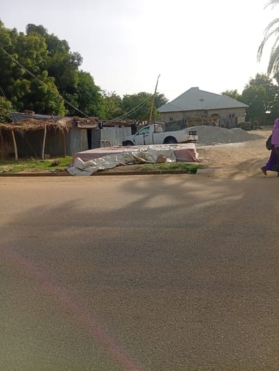 The image shows a street scene in Dutse, Nigeria, likely during the daytime given the bright, hazy sunlight. In the foreground, a paved road occupies the majority of the frame, with shadows cast across it, suggesting the sun is high.
To the left of the road, a small, rustic structure with a thatched roof and corrugated metal walls stands. In front of it, there are some green plants and a pile of white tarps or fabric. Behind these, a white pickup truck is parked, facing towards the right. Further back, a large, modern-looking house with a silver, sloped roof is visible. A significant pile of gravel or construction material is situated in front of this house, indicating ongoing construction or renovation.
On the right side of the image, a person dressed in a purple garment and a light pink headscarf is walking away from the camera, towards the right. A palm tree frond is visible at the very top right edge of the frame. The overall setting appears to be a mix of residential areas, with both simple and more substantial dwellings, and a rural or semi-urban atmosphere. The weather seems to be clear but hazy, possibly indicating warm temperatures. There is no visible text in the image.