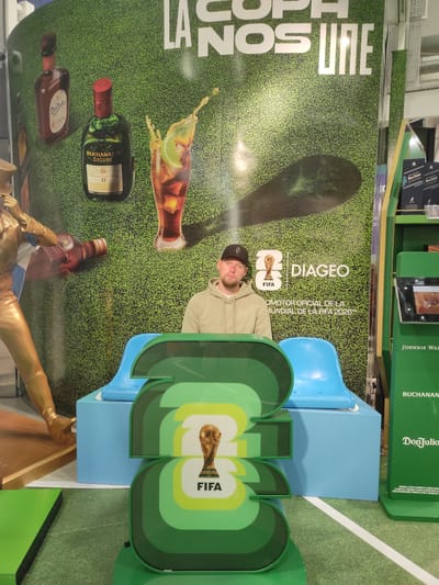 The image shows a man wearing a black baseball cap and a beige hoodie, seated in a blue molded plastic seat. He is positioned in front of a large backdrop featuring a grassy texture and graphics of liquor bottles, including Don Julio and Buchanan's Deluxe. The backdrop also displays the FIFA World Cup trophy and logos for Diageo and FIFA. To the right of the man, a green display stand holds bottles of Johnnie Walker and Buchanan's, along with product information. In the foreground, large green cutouts shaped like numbers or abstract forms are visible, also featuring the FIFA World Cup trophy and the FIFA logo. The scene appears to be a promotional display, likely at an event or retail location. The location is identified as the city of Ezeiza, Argentina.