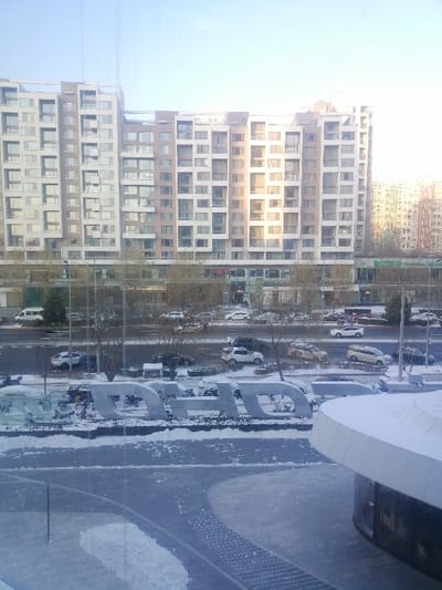 This image presents an elevated view of a snow-covered urban landscape in Fengtai District, China. In the background, a prominent multi-story building complex with a light-colored facade, featuring a grid of numerous rectangular windows, dominates the skyline. Sunlight reflects off parts of this structure.
In the mid-ground, a multi-lane asphalt road, partially cleared but still showing snow accumulation, is visible. Multiple vehicles, including sedans, SUVs, and a white van, are present on the road, some in transit and others parked. Bare deciduous trees, dusted with snow, line the road and are interspersed within small planting areas. Tall streetlights are positioned along the thoroughfare.
The immediate foreground features a large, snow-covered sculptural installation spelling out the letters "HD" in light-colored, block capitals. To the lower right, a modern, curved building or structure with a white roof is present, also partially covered in snow. The ground in the foreground consists of snow-covered paved areas and pathways. The sky above is bright and hazy. No human figures are discernible in the scene.