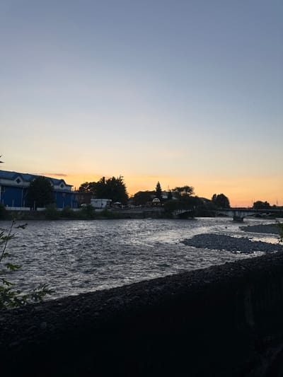 The image depicts a serene riverside scene during sunset, captured from a low angle. The sky transitions from a soft, pale blue to a warm gradient of orange and pink near the horizon, indicating the time of day is late evening. Wispy clouds are visible, adding texture to the sky.
The foreground is dominated by a fast-flowing river, its surface textured with ripples and waves that catch the fading light. The riverbank on the right is dark and rocky, transitioning into deep shadow. On the left bank, a few trees stand with their leaves dark against the twilight sky. Further back, a cluster of buildings with distinctive blue roofs and white accents are visible, nestled amongst greenery. Some of these buildings appear to be houses or small commercial establishments.
Beyond the buildings and trees, a bridge spans the river, with visible structural elements and what appear to be streetlights or lampposts. The scene suggests a settled area, perhaps a town or village, situated alongside the natural beauty of the river. The overall mood is tranquil and peaceful, evoking a sense of calm as day turns into night. There is no visible text in the image.