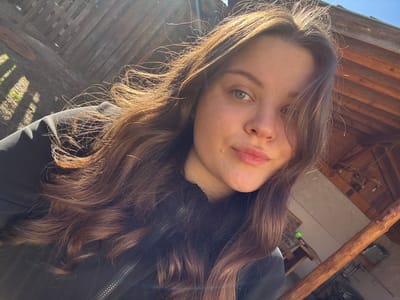 A young adult female is depicted in a close-up, upward-angled selfie, presumably taken outdoors in Raduil, Bulgaria. She has long, wavy, dark brown hair, back-lit by direct sunlight, creating a luminous effect. Her green eyes are open, and she exhibits a slight, closed-lip smile. A small mole is visible on her left cheek. She is wearing a black zippered jacket or hoodie. The background on the left features a weathered wooden fence with visible sunlight and shadows, along with patches of green vegetation. On the right, a wooden pergola or outdoor shelter structure is partially visible, characterized by prominent wooden beams and a lighter colored wall. A thick, reddish-brown wooden post extends diagonally upwards from the lower right corner of the frame. The scene is brightly illuminated by natural sunlight.