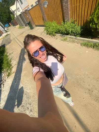 An outdoor, eye-level, medium selfie depicts a woman standing on a dirt road in an urban or suburban area. The woman has long, dark brown hair, wears oversized sunglasses, a light pink crop top, and light-wash jeans. She is sticking her tongue out and holding her hair with her left hand. A grey fanny pack is slung across her body. A long shadow of the woman is cast on the ground to her left, indicating the sun is behind her. The background shows a fence with vertical wooden planks, brick pillars, a green trash can, and houses with light-colored facades. There are also green bushes and trees visible. The overall environment suggests a residential street or alleyway within the city of Mosmetallokonstruktsiya Ont, Russia.